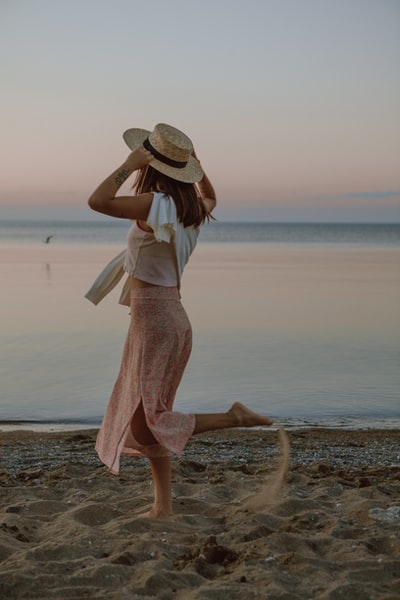 Standing on the beach during the day of a women wearing a white dress, wearing a brown hat
